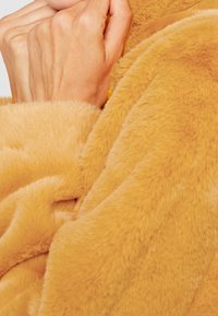 Händerna hopknutna och vilar på ett mjukt, ljusbrunt, lurvigt tyg, vilket föreslår värme och komfort.