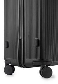 Valise rigide noire avec une surface texturée, fermeture éclair et quatre roues arrondies pour la mobilité. Équipée de poignées de transport encastrées.