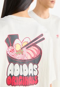 White cotton t-shirt featuring a colorful ramen bowl graphic on the back. The design includes noodles, eggs, and chopsticks, with bold "ADIDAS ORIGINALS" text.