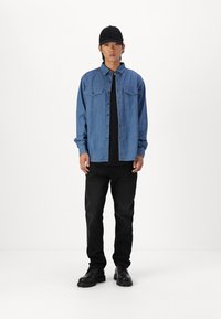 Camicia in denim di colore azzurro chiaro, maniche lunghe, con tasche a scatto frontali, abbinata a una T-shirt nera e jeans scuri, completata da stivali neri e un berretto.