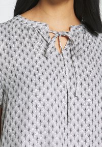 White blouse with black geometric pattern, slightly gathered neckline, tied keyhole opening, and short sleeves. Soft, lightweight fabric.