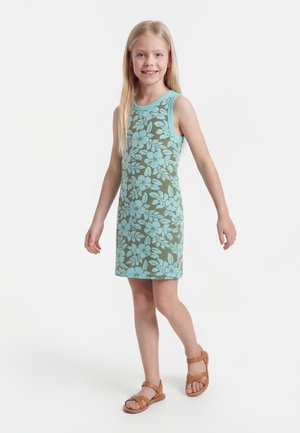 Dziewczyna stoi i uśmiecha się, ubrana w bezrękawnikową zieloną sukienkę z niebieskimi kwiatowymi wzorami i brązowe sandały na białym tle.