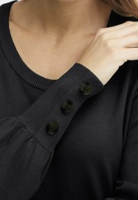 Svart stickad tröja med rund halsringning och puffärmar, med tre glänsande svarta knappar på ärmslutet för detalj.