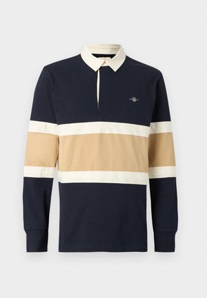 Ragbi majica z dolgimi rokavi v mornarsko modri, bež in beli črtasti vzorec, s kremnim ovratnikom in majhnim izvezenim logotipom na levi strani prsi.