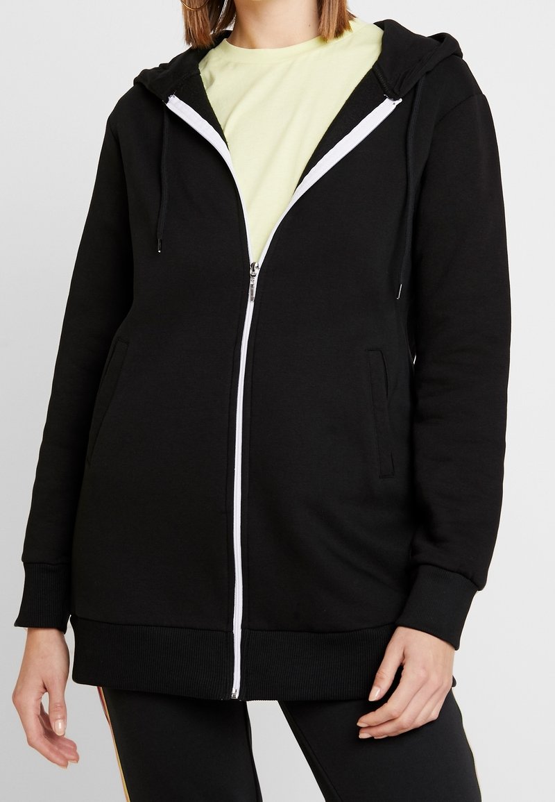 Personne portant un sweat à capuche noir zippé avec une fermeture éclair blanche et un t-shirt jaune clair en dessous, posant devant un fond uni.