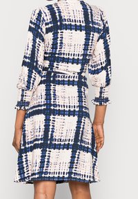 Femme portant une robe mi-longue à motif à carreaux bleu et blanc, manches 3/4 bouffantes et poignets smockés, vue de dos.