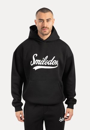 Zwarte hoodie gemaakt van een katoenmix, met een witte "Smilodox" logo, kangoeroezak en geribbelde manchetten. Gecombineerd met bijpassende zwarte broeken.
