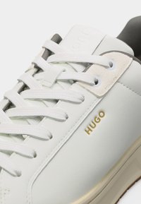 Κοντινή λήψη λευκού αθλητικού παπουτσιού με λευκά κορδόνια, μπεζ τρύπες, μαύρη εσωτερική επένδυση, μπεζ σόλα και χρυσό λογότυπο "HUGO" στη πλευρά.