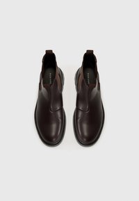 Zapatos marrones de piel deslizantes con punta redondeada, superficie texturizada, acentos laterales elásticos y suela de goma estriada. Logo de la marca en la plantilla.