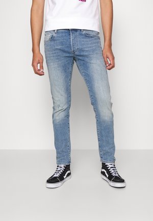 Jeans ajustados de ligero azul con textura desgastada y un ligero desgarro, combinados con zapatillas altas negras y una camiseta blanca.