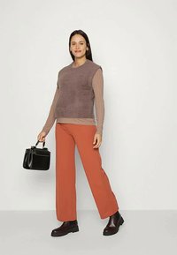 Mulher de pé, vestindo um sweater castanho sem mangas sobre uma blusa de mangas longas, calças cor ferrugem, botas escuras e segurando uma bolsa preta.