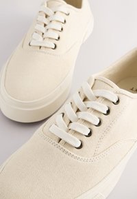 Smėlio spalvos drobiniai sportbačiai su tekstūruotu paviršiumi, balti batraiščių raiščiai ir juodi akių žiedeliai. Apvalios noselės ir lygi guminė pado dalis.