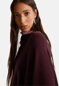 Giovane donna con lunghi capelli castani che indossa orecchini a cerchio d'argento, un maglione di maglia bordeaux e un dolcevita viola chiaro su uno sfondo bianco.