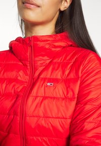 Donna che indossa una giacca trapuntata rossa brillante con un piccolo logo rettangolare sul petto e lunghi capelli scuri visibili sulla spalla.
