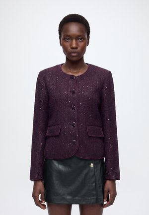 Femme portant une veste boutonnée violet foncé avec des poches et une jupe en cuir noire, debout devant un fond blanc uni.
