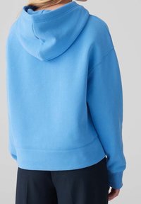 Osoba oblečená v volné světle modré mikině s kapucí, v kombinaci s černými kalhotami, zobrazená zezadu na neutrálním pozadí.