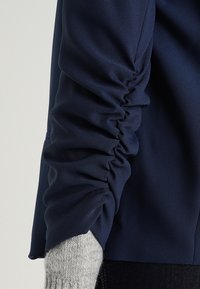 Blusa azul marino con detalle de mangas recogidas, hecha de una tela suave. Puño de punto gris visible debajo, que añade contraste de textura.