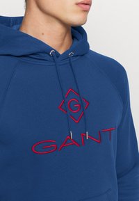 Sudadera azul real hecha de tejido suave, con un gran logotipo rojo bordado. Cordoneras con extremos de metal y mangas raglán.