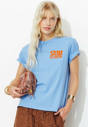 Femme blonde aux cheveux ondulés porte un t-shirt bleu "SUN LOVER", une jupe en dentelle marron, des bijoux en or, et tient une pochette marron à motifs.