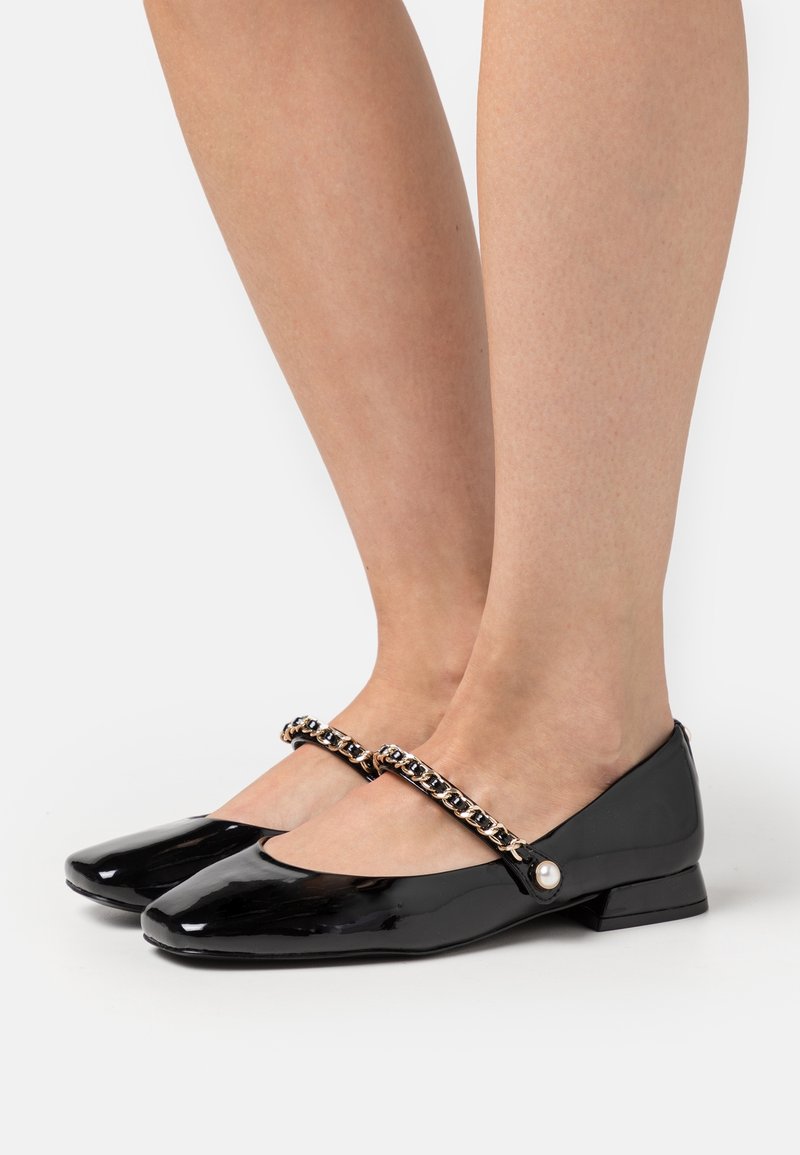 Chaussures plates Mary Jane en cuir verni noir avec brides en chaîne et détail bouton perle, portées pieds nus.