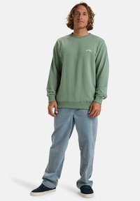 Mintgrüner Crewneck-Sweatshirt mit gerippten Bündchen und Saum. Mit kleinem weißen Logo auf der Brust. Kombiniert mit hellblauen Denim-Hosen und dunklen Sneakers.