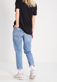 Hellblaue Denim-Jeans, lockere Passform, mit umgeschlagenen Säumen, kombiniert mit einem schwarzen T-Shirt und weißen Turnschuhen, stehen vor einem schlichten Hintergrund.