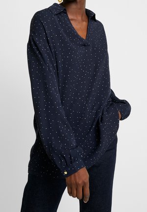 Femme portant un chemisier manches longues bleu marine à petits pois blancs et poignets à boutons dorés, associée à un pantalon foncé, main dans la poche.