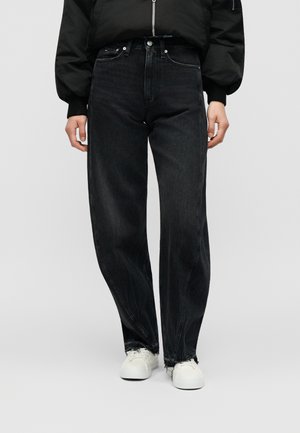 Άτομο που φοράει μαύρα τζιν με ψηλή μέση και φαρδιά μπατζάκια με ξεφτισμένες άκρες, μαύρο μπουφάν και λευκά αθλητικά παπούτσια, στέκεται μπροστά σε μια απλή φόντο.