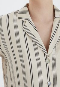 Camicia a maniche corte, con bottoni, di colore beige con sottili righe verticali nere. Presenta un colletto a punta e bottoni rotondi con una leggera lucentezza.