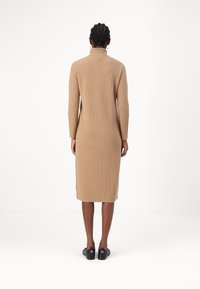 Robe en maille couleur camel avec un col montant, des manches longues et une coupe droite, s'étendant sous le genou avec des fentes latérales.