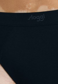 Černé bavlněné boxerky s elastickým pasem s logem "sloggi", hladká textura a minimalistický design. Detailní záběr na látku.