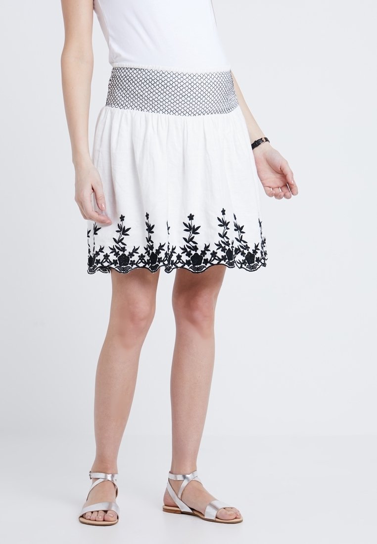 Jupe blanche avec broderie florale noire à l'ourlet, une taille en tissu texturé noir et une coupe évasée. Associée à des sandales argentées.