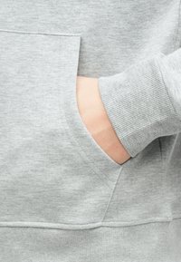 Main partiellement dans la poche avant d'un sweat-shirt gris clair avec poignets côtelés et texture de tissu douce.