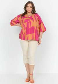 Kleurrijke oversized top in oranje en roze abstracte patronen, gecombineerd met lichtbeige cropped pants en oranje sandalen met banden.