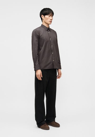 Άνδρας που φοράει σκούρο γκρι πουκάμισο με κουμπιά, μαύρο παντελόνι φαρδιάς γραμμής και καφέ αθλητικά παπούτσια, στέκεται μπροστά σε απλό λευκό φόντο.