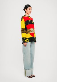 Pleteni pulover v rdeči, rumeni in črni barvi z abstraktnimi vzorci; v kombinaciji s svetlo modrimi ohlapnimi jeans hlačami in črnimi sandali.