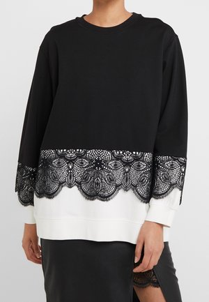 Kvinde iført en sort og hvid top med lange ærmer og sort blonderkant ved kant og manchetter, kombineret med en sort nederdel med blonderdetalje.