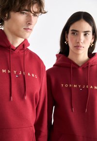 Sudaderas rojas de tela suave, con el texto dorado "TOMMYJEANS" y pequeños acentos de logo. Los cordones añaden funcionalidad al diseño.
