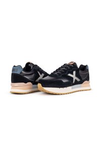 Schwarze Wildleder- und Lederschuhe mit einer klobigen Sohle, die ein auffälliges weißes "X"-Logo und hellblaue Akzente an der Ferse aufweisen.