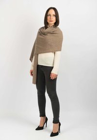 Beige sjal lavet af blødt stof, draperet omkring skuldrene. Modellen bærer en lys sweater og mørke skinny jeans, med sorte højhælede sko.