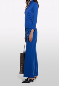 Blå långärmad topp med hög krage, matchad med vida blå byxor. Modellen bär en grå tote-väska och svarta mule-skor.