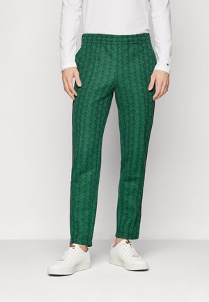 Man draagt groene slim-fit broek met geometrisch patroon, witte blouse met lange mouwen en witte sneakers, staande tegen een effen achtergrond.
