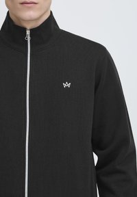 Veste zippée noire avec un col haut, texture de tissu lisse, et un petit logo brodé blanc sur la poitrine.