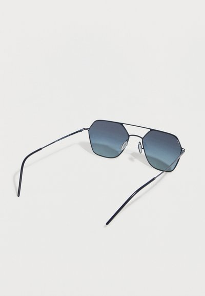 Μαύρα λεπτά γυαλιά ηλίου με μεταλλικό σκελετό και γεωμετρικούς φακούς με μπλε βαθμιαία απόχρωση, καθώς και ρυθμιζόμενα μαξιλαράκια μύτης σε λευκό φόντο.