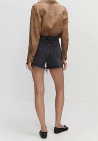 Shorts de mezclilla con dobladillo deshilachado, color gris oscuro, combinados con una camisa de botones marrón, costuras visibles y bolsillos. Zapatos planos negros.