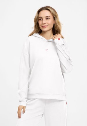 Woman wearing white hoodie and matching white pants, with a small red logo on the chest and sleeve, standing against a white background.