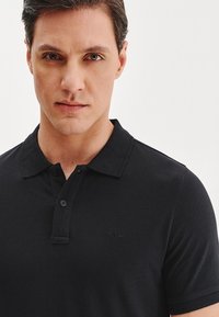 Czarny polo z klasycznym kołnierzykiem, trzema guzikami i subtelną teksturą. Logo marki umieszczone na piersi.