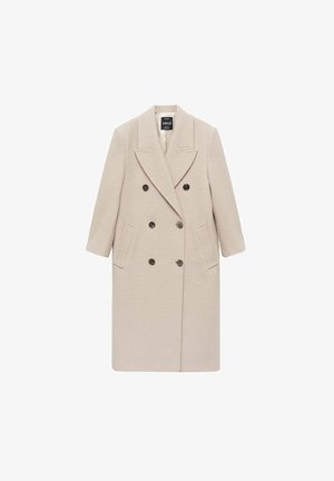 Persona que lleva un abrigo cruzado de lana beige, suéter de canalé crema, camisa blanca con cuello y un cinturón negro fino en la cintura.
