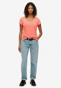 Vrouw met krullend haar in een koraalkleurig t-shirt, lichte spijkerbroek, zwarte riem en zwarte schoenen, met één hand in haar zak.