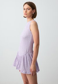 Lila jurk van textuurstof, mouwloos ontwerp en uitlopende rok. Heeft een aansluitend lijfje en een gerimpelde zoom. Gladde oppervlakte.